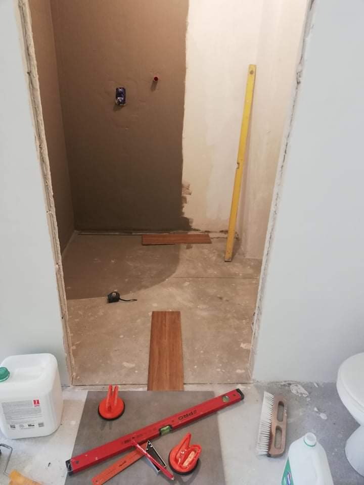 Łazienka w trakcie remontu: częściowo położone płytki podłogowe imitujące drewno, na podłodze poziomica, przyssawki do płytek i wiadro z klejem. Ściana częściowo pokryta szarą masą, widoczne...