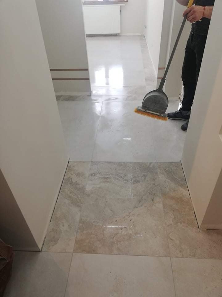 Układanie jasnych, marmurkowych płytek podłogowych w korytarzu, widoczny pracownik sprzątający szczotką z szufelką, przejście do kolejnego pomieszczenia z białymi ścianami i jasną podłogą.