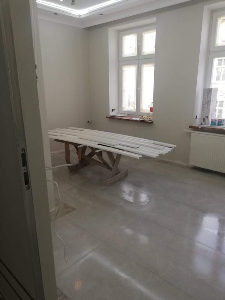 Przygotowana do malowania listwa przypodłogowa leży na drewnianym stole w pokoju z dwoma oknami, po remoncie, z nowymi płytkami podłogowymi i świeżo pomalowanymi ścianami na biało.