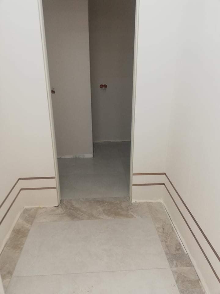 Wnętrze w trakcie wykańczania: białe ściany z dekoracyjnymi brązowymi paskami przy podłodze, jasna podłoga z płytek imitujących marmur, widoczne surowe ściany w przejściu do innego pomieszczenia.