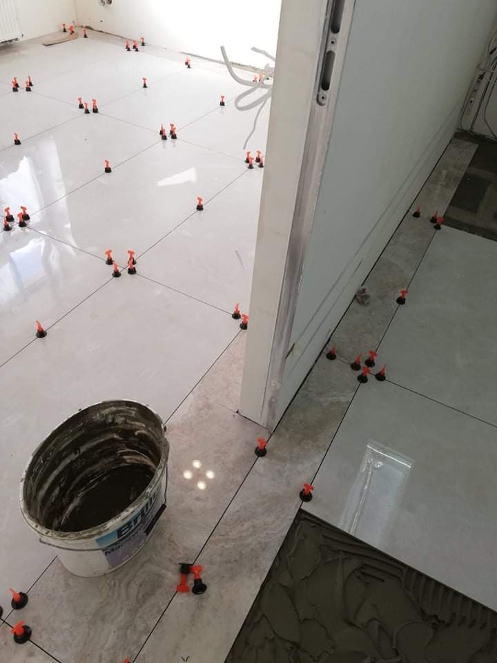 Proces układania jasnych, błyszczących płytek podłogowych z użyciem systemu poziomowania płytek (klipsy i kliny); widoczne wiadro z klejem i świeżo nałożony klej na płytce.