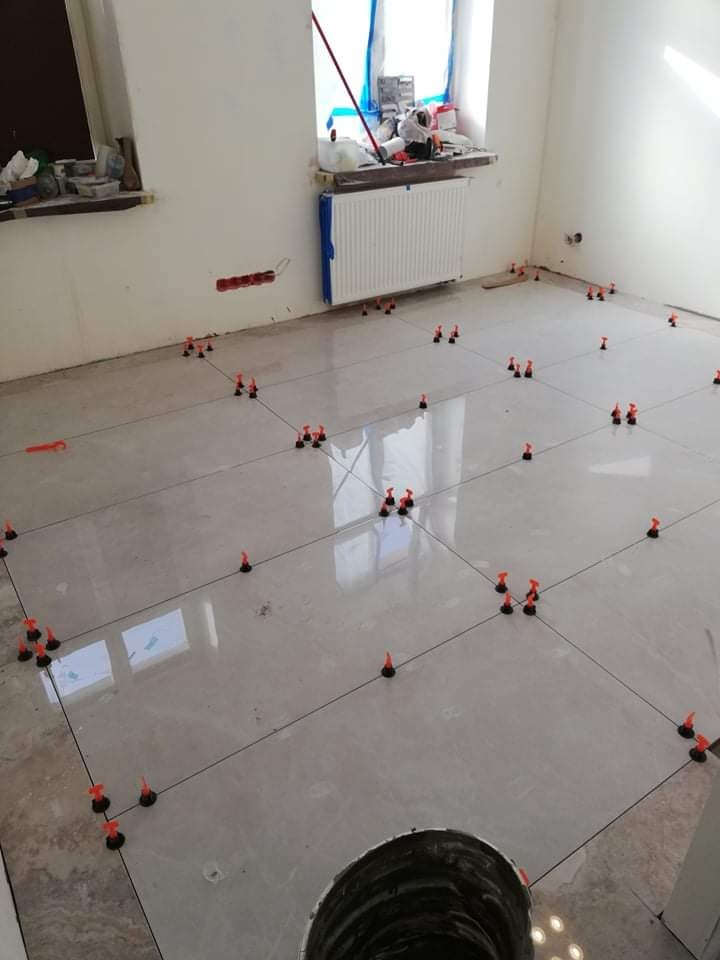 Układanie płytek podłogowych z użyciem systemu poziomowania płytek w jasnym pomieszczeniu z grzejnikiem i oknem.