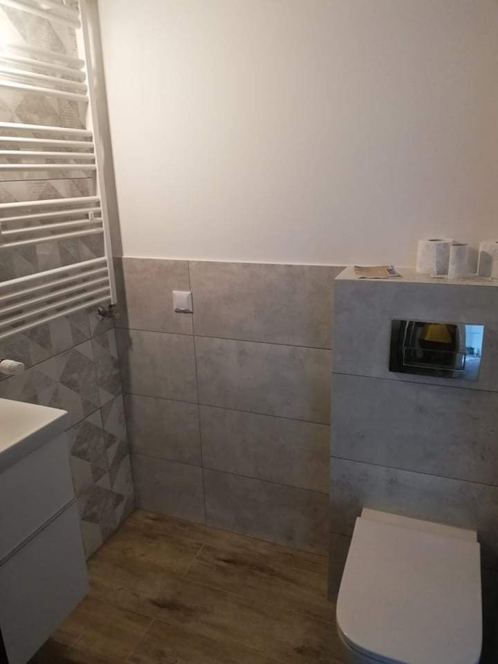 Wykończona łazienka z szarymi płytkami w stylu betonu, drewnopodobną podłogą i grzejnikiem drabinkowym, widok na umywalkę z szafką i toaletę w zabudowie.