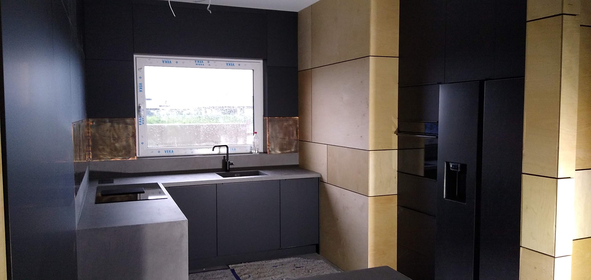 Nowoczesna kuchnia z ciemnymi, matowymi frontami szafek i blatem z betonu architektonicznego, akcentowana miedzianymi panelami nad blatem przy oknie oraz jasną drewnianą okładziną ściany...