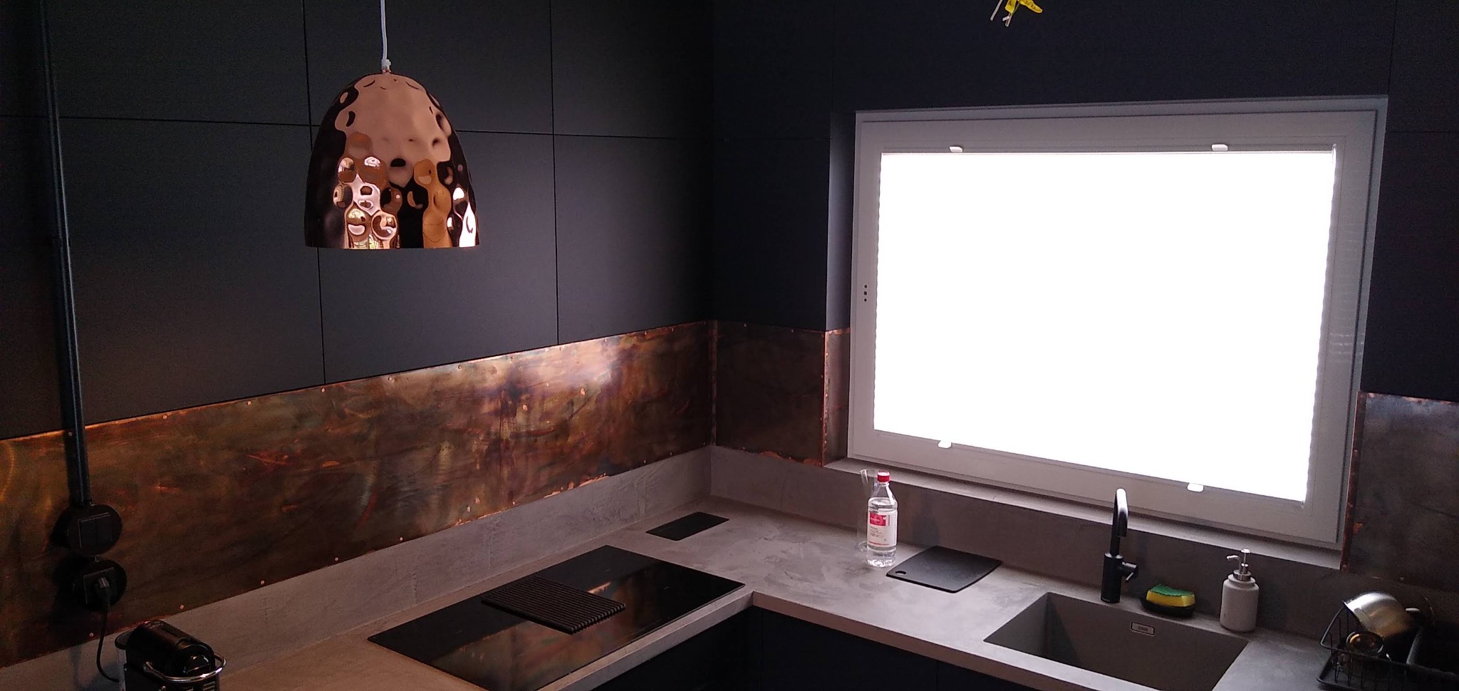 Nowoczesna kuchnia z ciemnymi szafkami i miedzianymi akcentami w Lublinie. Widoczny miedziany panel nad blatem, designerska lampa i okno z białymi ramami.