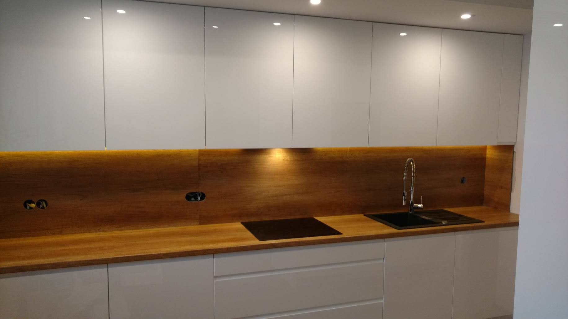 Nowoczesna kuchnia z białymi, lakierowanymi szafkami górnymi i dolnymi, drewnianym blatem i panelem ściennym z ukrytym oświetleniem LED, czarny zlew i płyta indukcyjna.
