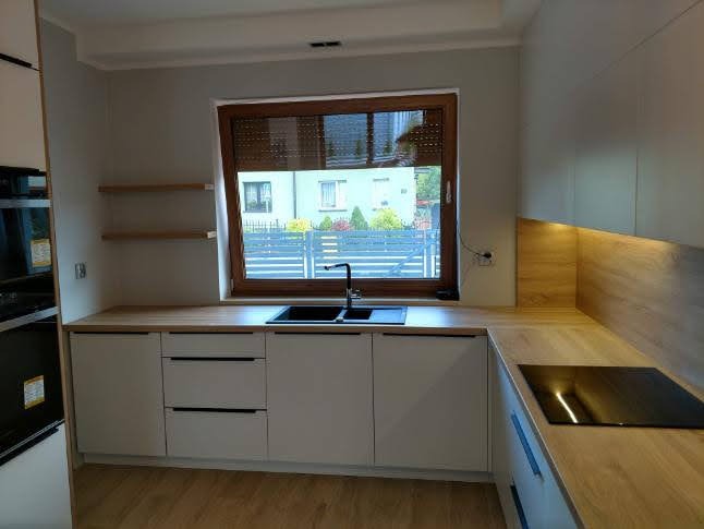 Nowoczesna kuchnia z białymi frontami, drewnianym blatem i oknem z widokiem na osiedle domów jednorodzinnych.