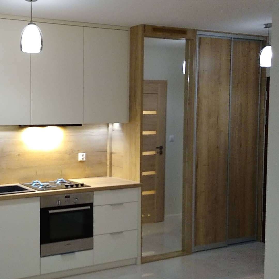 Nowoczesna kuchnia z białymi szafkami, drewnianym blatem i panelem ściennym imitującym drewno; obok zabudowana szafa przesuwna z lustrem, odbijająca drzwi wewnętrzne.