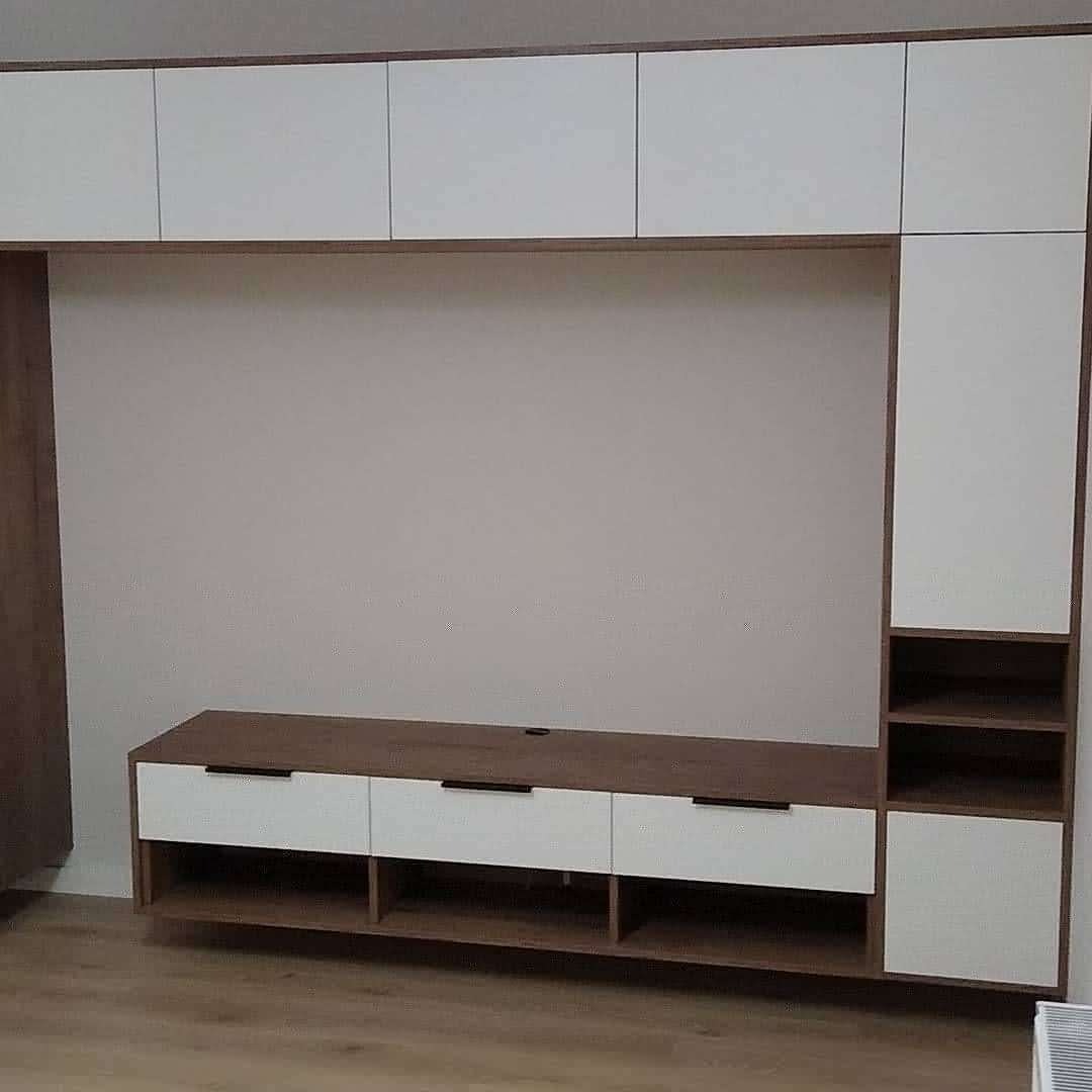 Zabudowa ściany z szafkami wiszącymi i szafką RTV z szufladami w kolorze białym i drewnopodobnym, z czarnymi uchwytami, na tle gładkiej ściany.