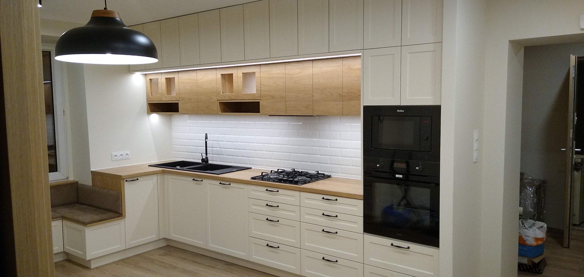 Jasna kuchnia z zabudową na wymiar, kremowe fronty szafek z czarnymi uchwytami, drewniany blat, czarny zlewozmywak i płyta gazowa, białe kafelki na ścianie, czarny piekarnik i mikrofalówka Amica...