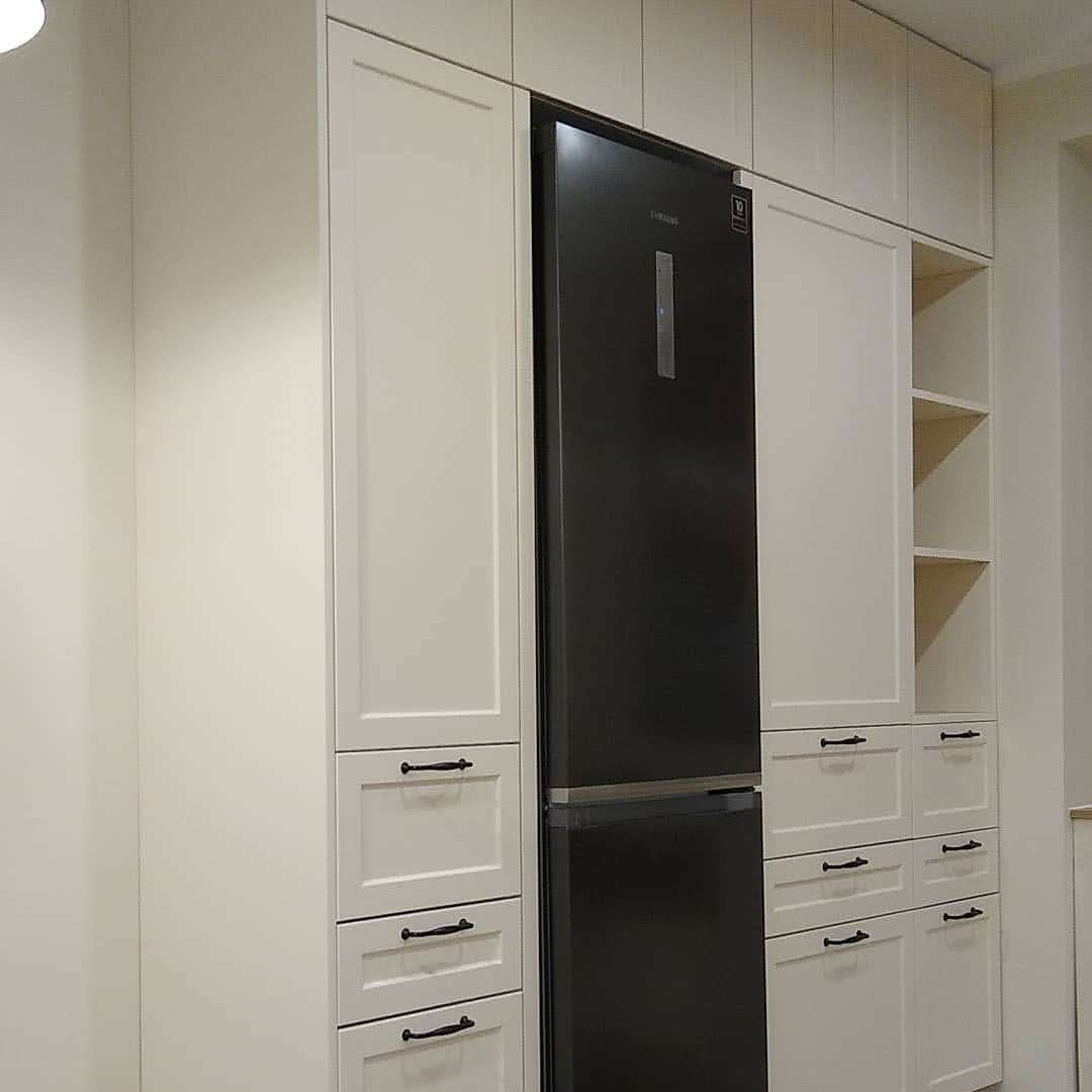 Biała zabudowa kuchenna z czarną lodówką wkomponowaną między szafki, widoczne minimalistyczne uchwyty i otwarte półki po prawej stronie.