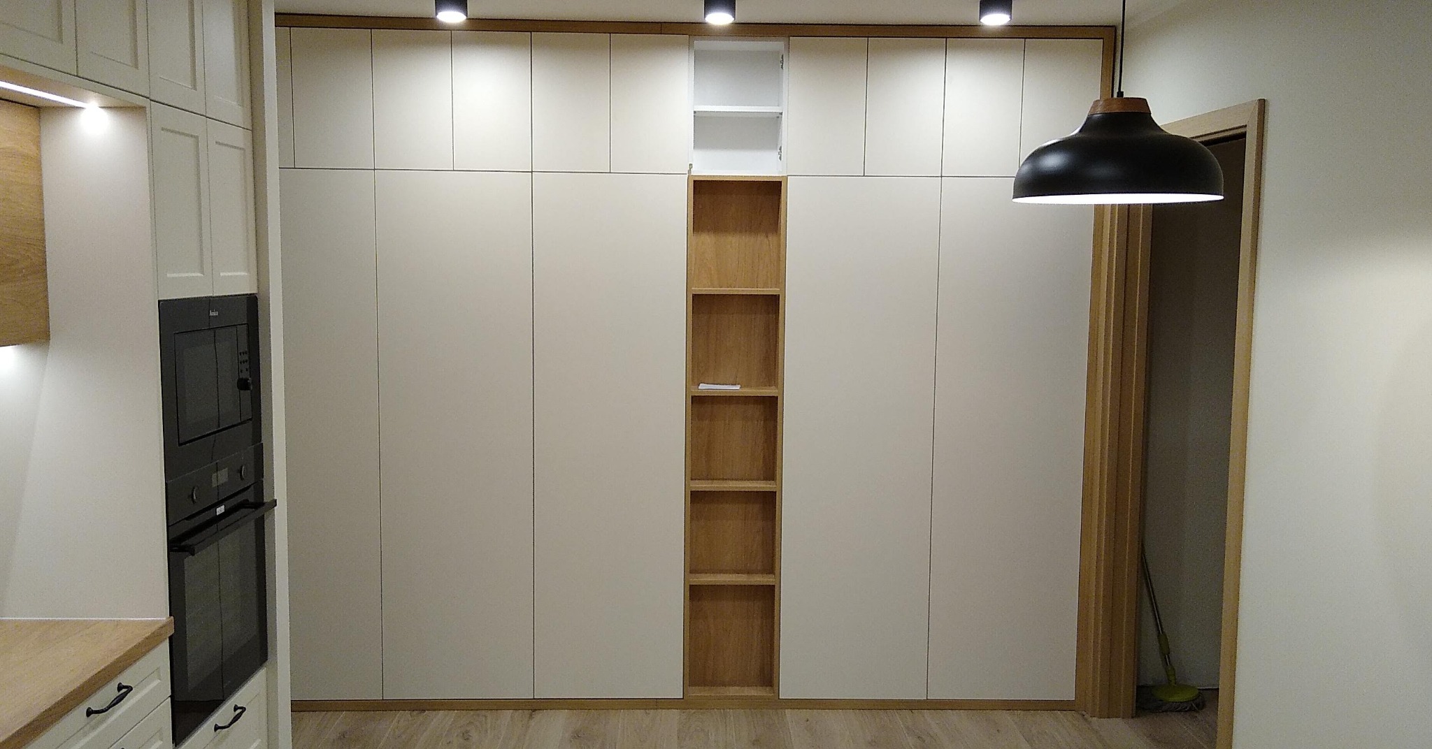 Duża, zabudowana szafa z białymi frontami i drewnianym regałem w środkowej części, zintegrowana z zabudową kuchenną, oświetlenie punktowe na suficie, podłoga z paneli.