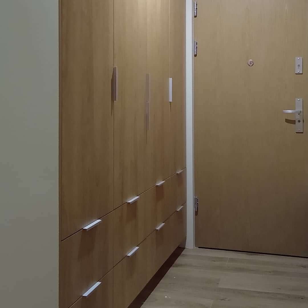Wysoka, drewniana szafa z szufladami i drzwiami, z minimalistycznymi, podłużnymi uchwytami, przylegająca do ściany obok drewnianych drzwi wejściowych.