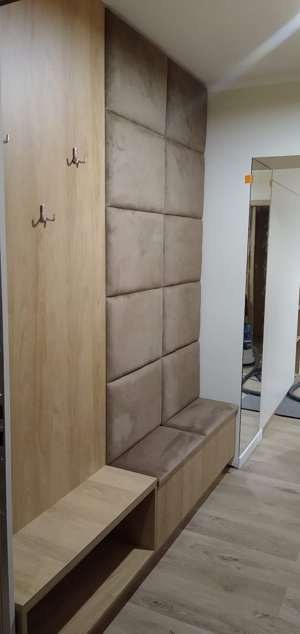 Wnęka z szafą z jasnego drewna z haczykami, tapicerowanym panelem ściennym w kolorze beżowym i siedziskiem oraz półką, widok na przedpokój z lustrem.