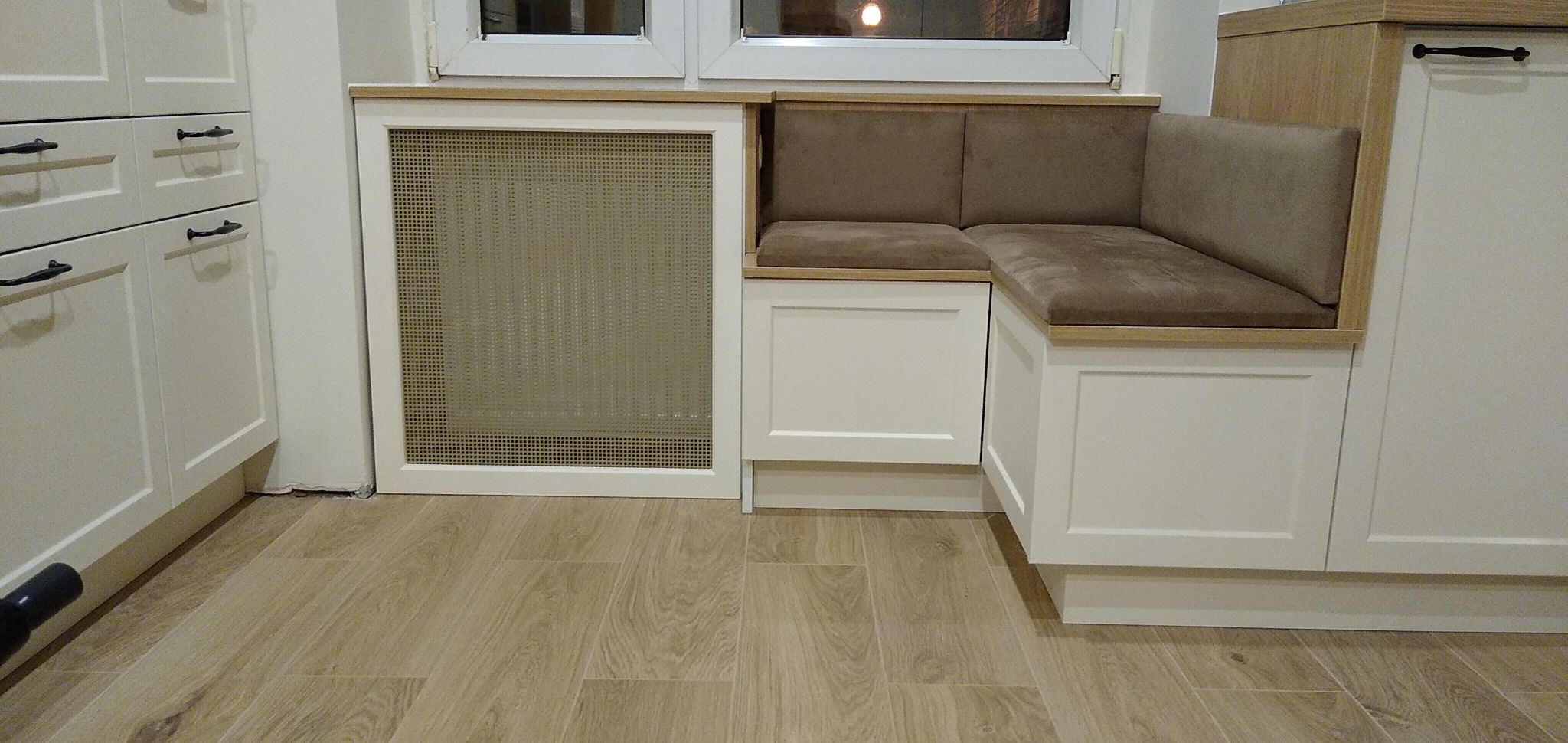 Biała zabudowa kuchenna z siedziskiem narożnym pod oknem, z tapicerowanymi poduchami w odcieniach brązu i obudowanym grzejnikiem, na podłodze panele imitujące drewno.