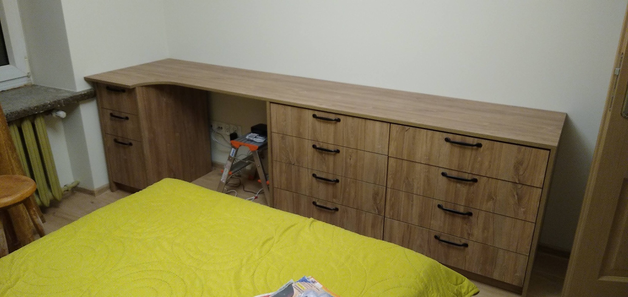 Długi, drewnopodobny blat biurka połączony z szafkami z szufladami i małą szafką, w tle drabina i gniazdka elektryczne, widoczne fragmenty pokoju z żółtą narzutą na łóżku.
