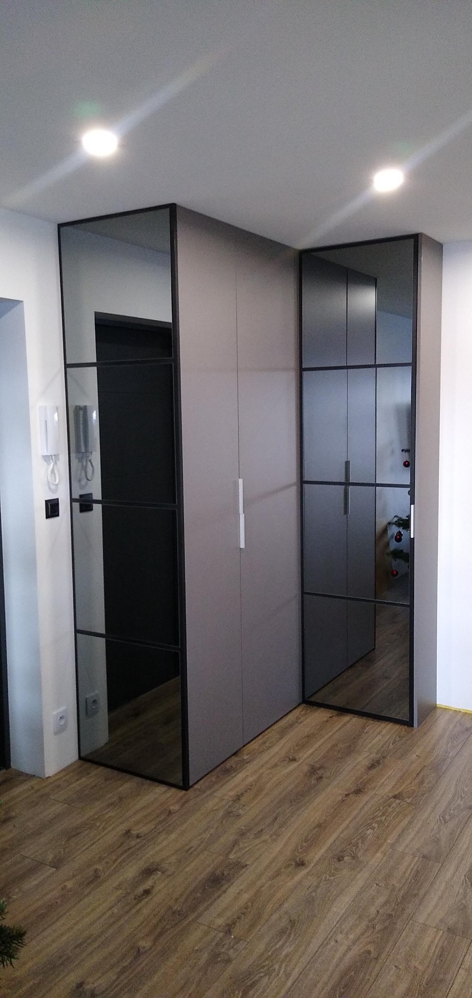 Szara szafa narożna z lustrzanymi drzwiami i czarnymi ramami, widoczna w przedpokoju z drewnianą podłogą i białymi ścianami.