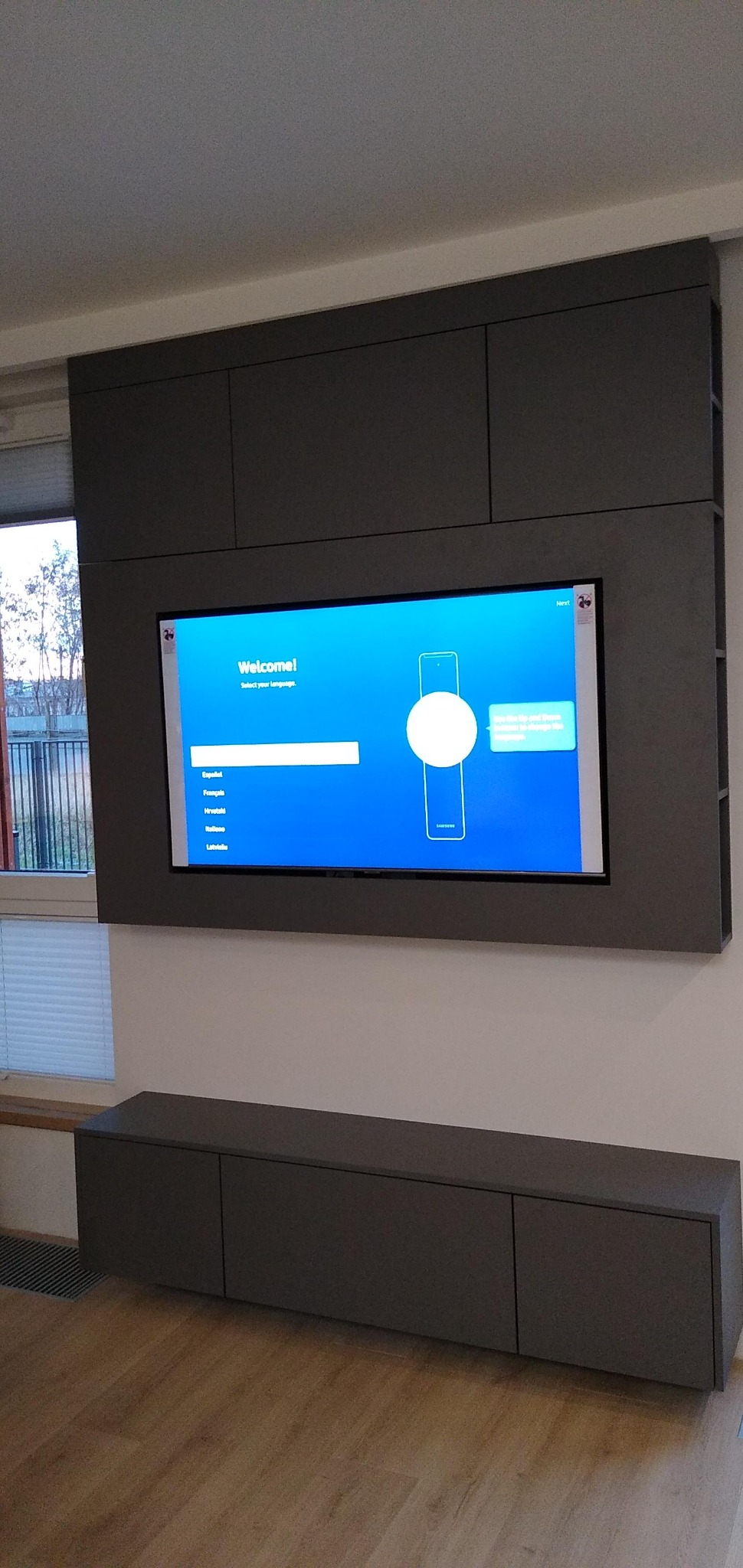 Szara zabudowa meblowa pod telewizor z szafkami wiszącymi i stojącymi, minimalistyczny design, widok na okno z roletami i drzewa za oknem.