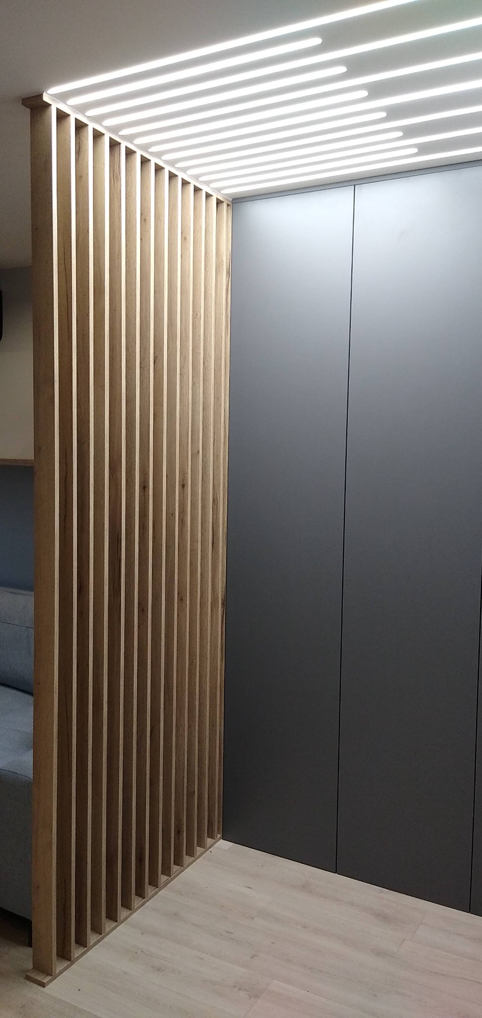 Drewniana ażurowa ścianka działowa z podświetleniem LED na suficie, obok szara zabudowa meblowa, jasna podłoga.