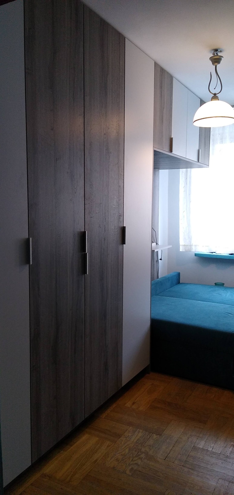 Wysoka szafa z frontami w kolorze drewna i białymi bokami, zintegrowana z zabudową pokoju, widoczny fragment niebieskiej sofy i parkiet na podłodze.