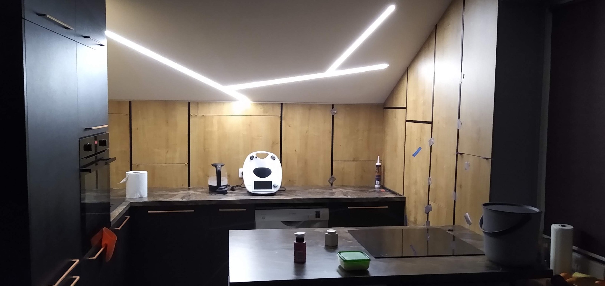 Montaż kuchni na poddaszu: widoczne panele ścienne z jasnego drewna, czarne szafki, nietypowe oświetlenie sufitowe LED w kształcie rozgałęzionych linii, silikon budowlany i taśmy maskujące wskazują...