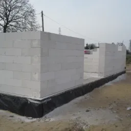 Wznoszenie ścian parterowego domu z bloczków betonu komórkowego, widoczna hydroizolacja fundamentów, teren budowy w pochmurny dzień.