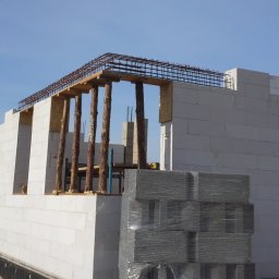 YTbud phu MONIC - Wznoszenie ścian budynku z betonu komórkowego, widoczne zbrojenie i drewniane stemple podtrzymujące konstrukcję stropu, paleta bloczków zapakowana w folię.