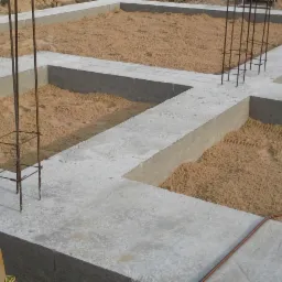 Zalane betonem fundamenty z wystającymi prętami zbrojeniowymi, wypełnione piaskiem, widoczne ślady opon w piasku.