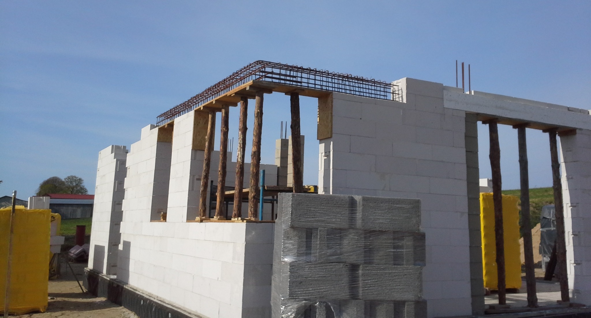 Wznoszenie ścian budynku z betonu komórkowego, widoczne zbrojenie i drewniane stemple podtrzymujące konstrukcję stropu, paleta bloczków zapakowana w folię.