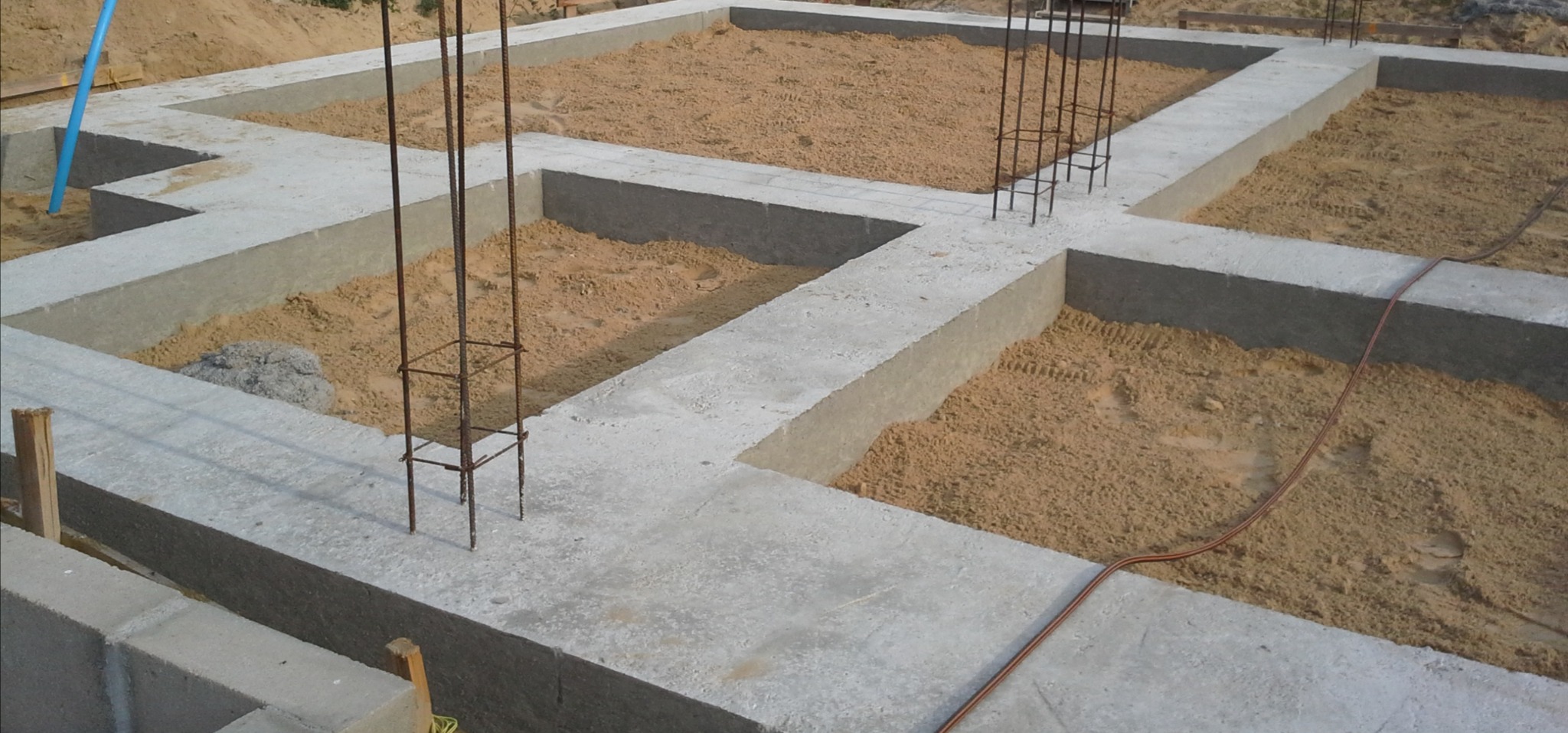 Zalane betonem fundamenty z wystającymi prętami zbrojeniowymi, wypełnione piaskiem, widoczne ślady opon w piasku.