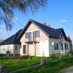 Dom w trakcie budowy z nowym, ciemnym dachem z blachodachówki, widoczne niezakończone ocieplenie elewacji z białego styropianu, brązowe ramy okienne i ceglany komin.