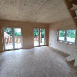 Wnętrze surowego domu z drewnianą konstrukcją, widoczne ściany i sufit z desek, podłoga z płyt OSB, okna z widokiem na zieleń i częściowo zmontowane drewniane schody.