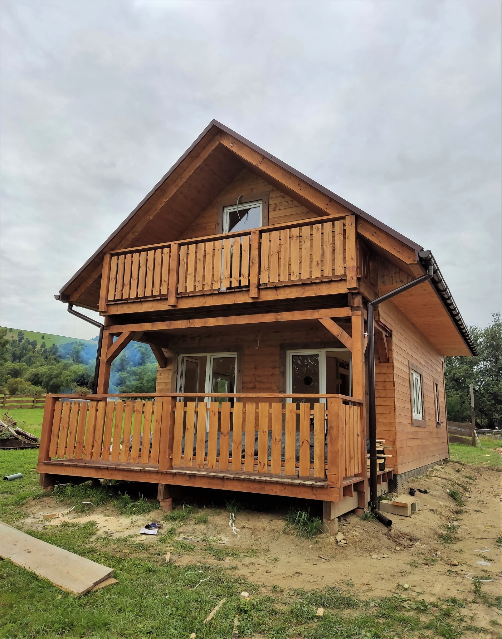 Dwukondygnacyjny, drewniany dom z balkonami, widok z zewnątrz na tle zalesionych wzgórz i pochmurnego nieba.