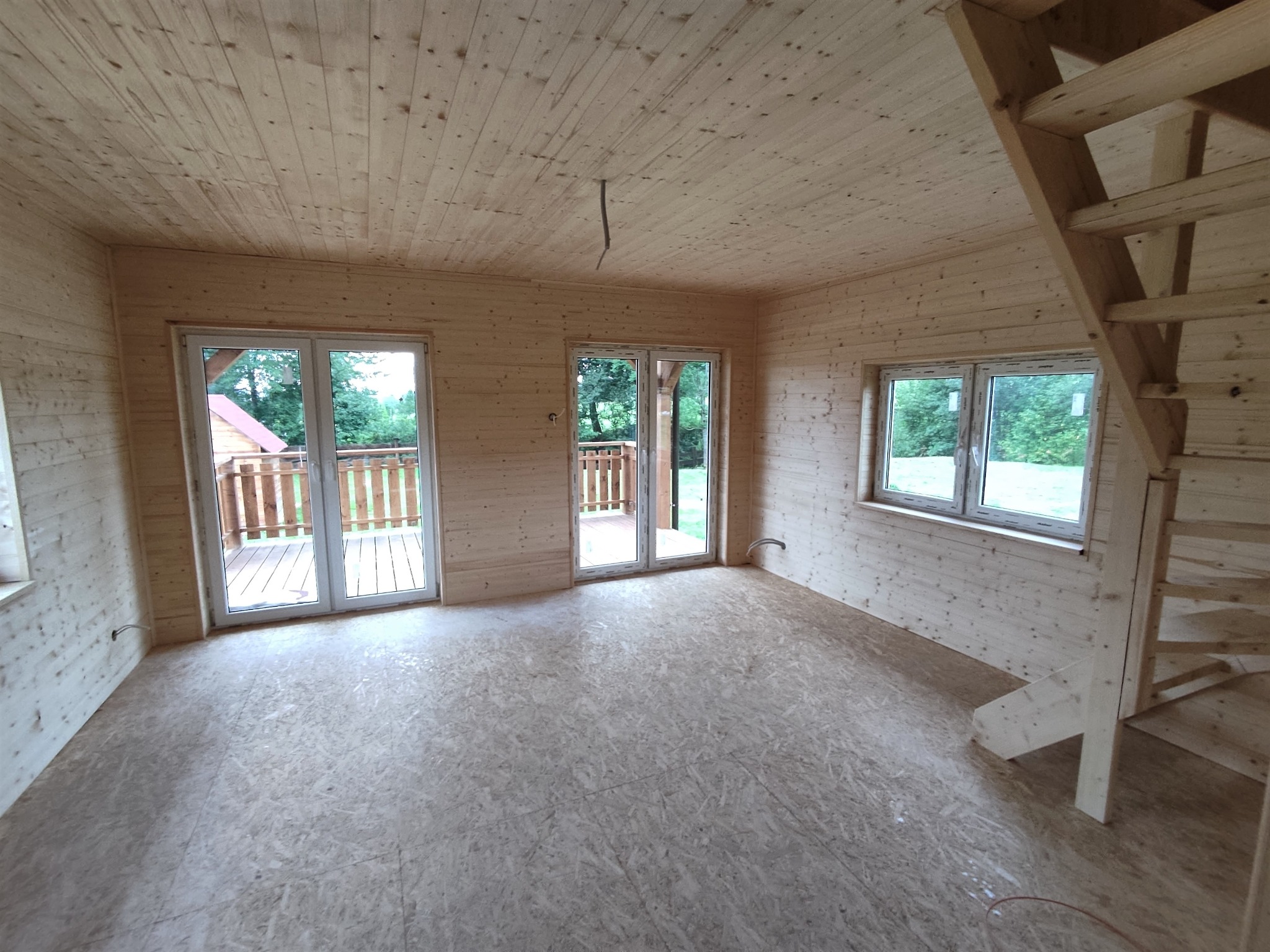 Wnętrze surowego domu z drewnianą konstrukcją, widoczne ściany i sufit z desek, podłoga z płyt OSB, okna z widokiem na zieleń i częściowo zmontowane drewniane schody.