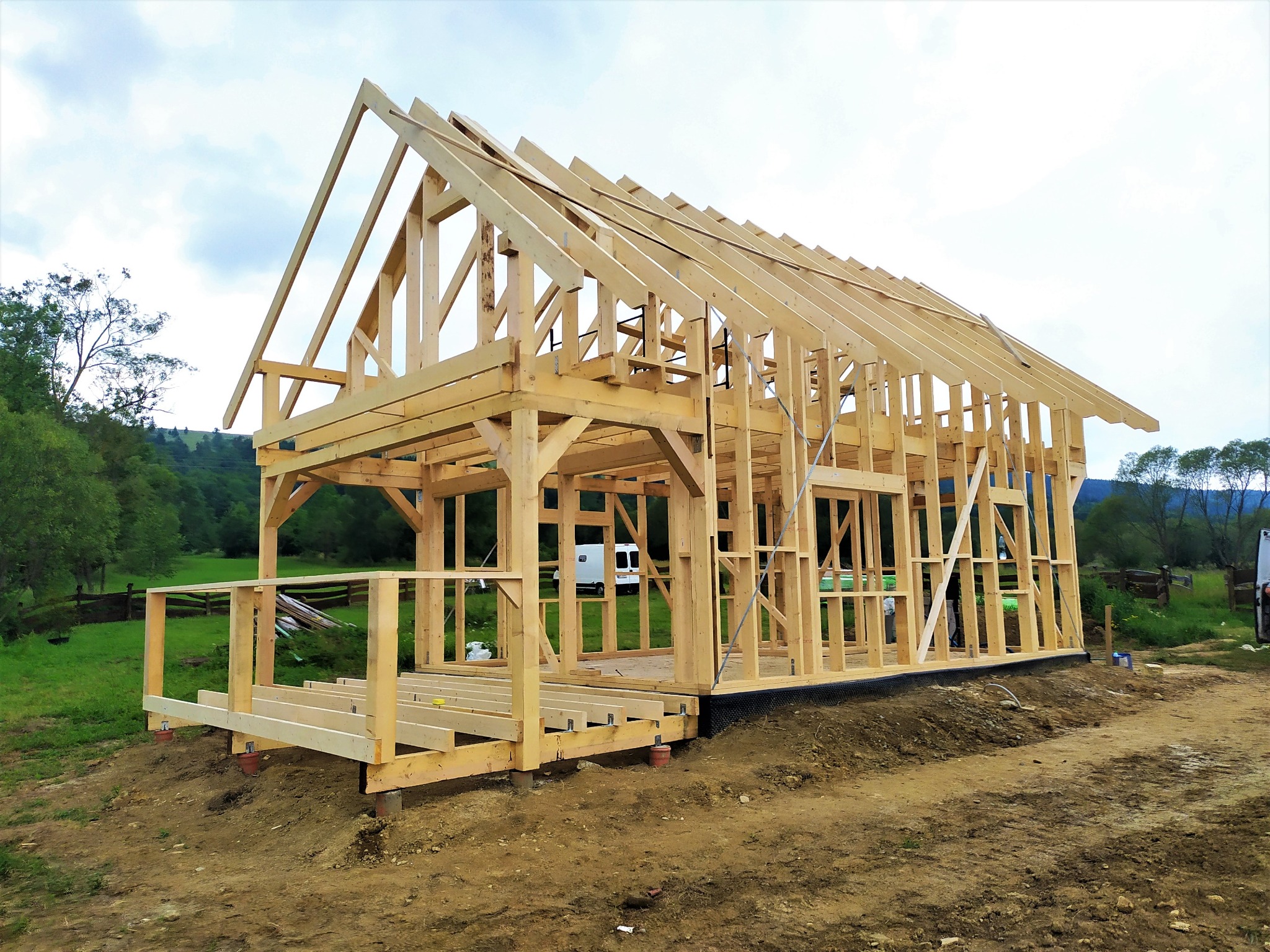 Wznoszenie konstrukcji domu z drewna, widoczne elementy szkieletu, krokwie i słupy, w tle zieleń i niebo z lekkimi chmurami.