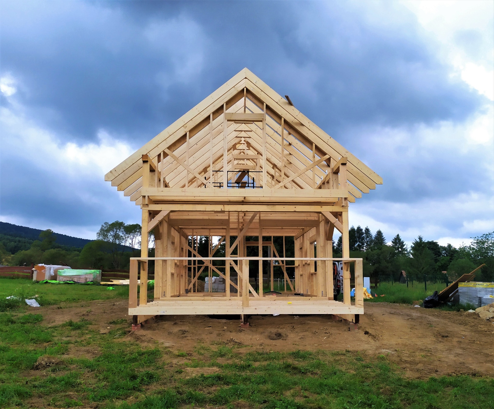 Surowy szkielet drewniany domu w trakcie budowy, widok z przodu na tle pochmurnego nieba i zieleni