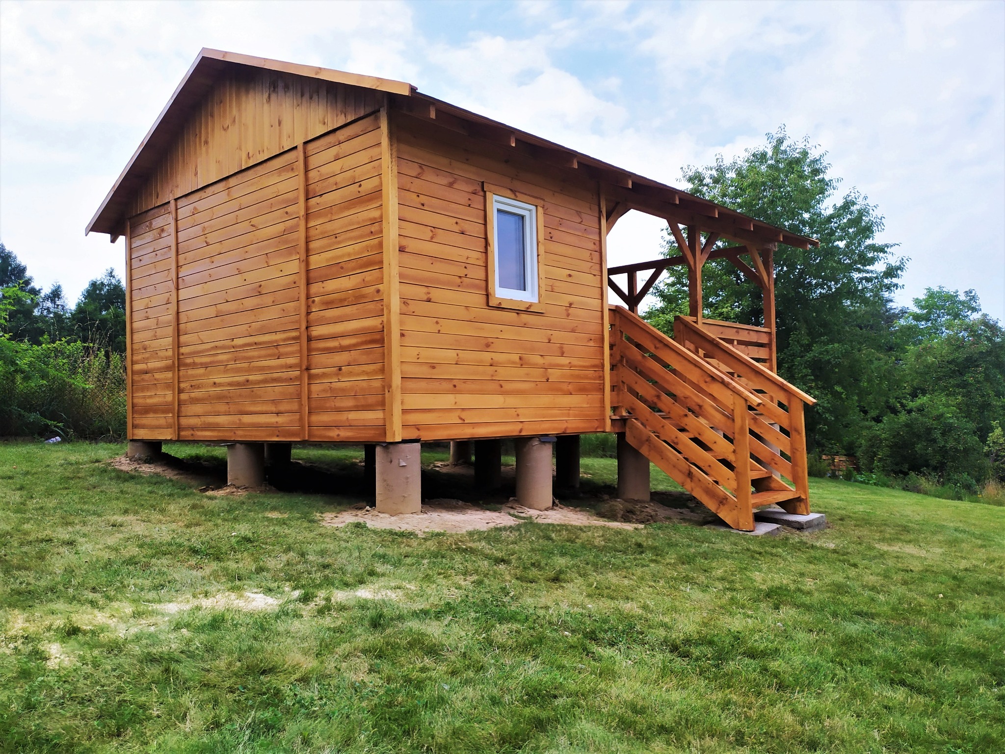 Niewielki drewniany domek letniskowy, wzniesiony na betonowych słupach, z drewnianymi schodami i zadaszonym tarasem.