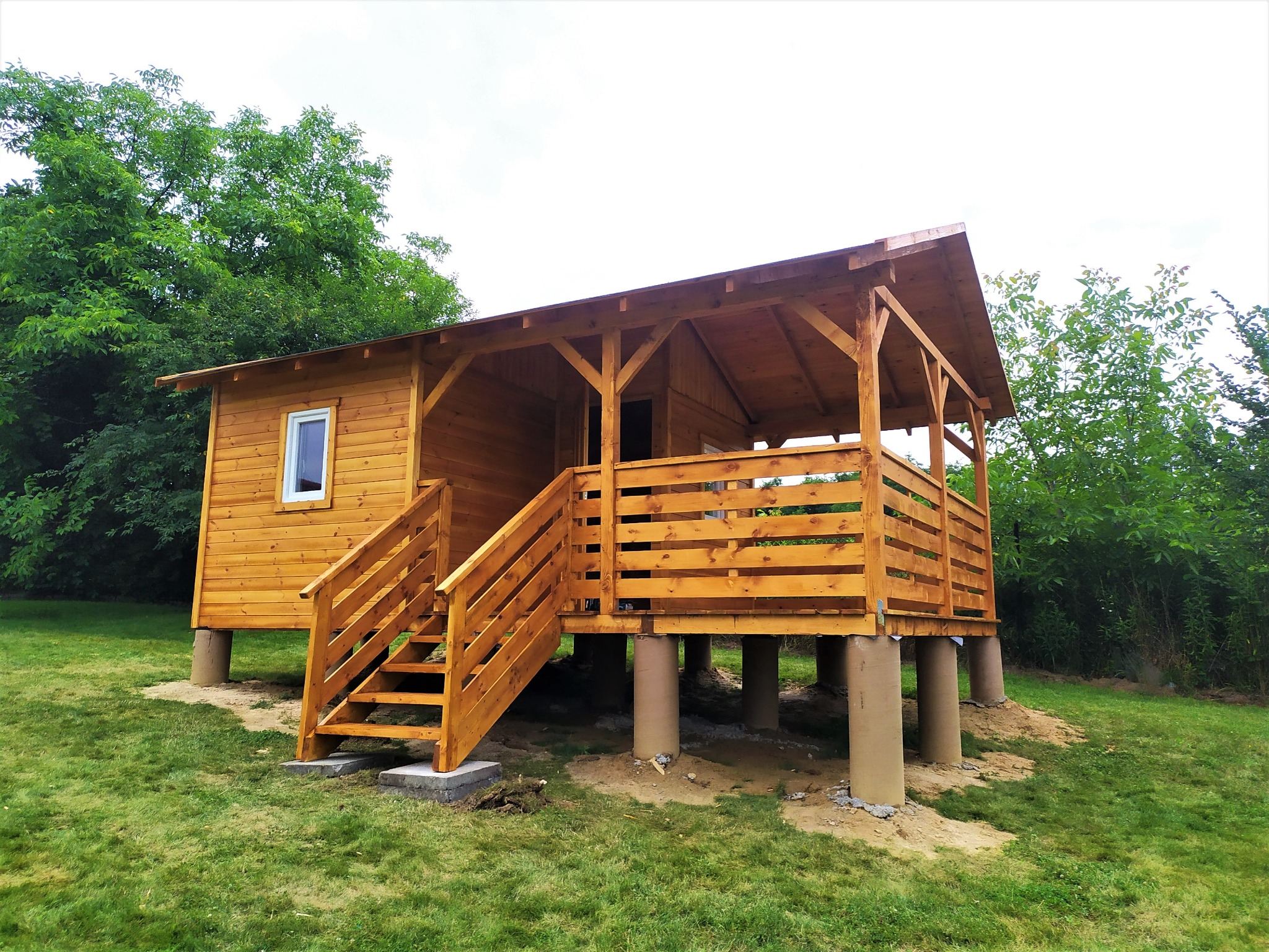 Niewielki drewniany domek letniskowy na podwyższeniu z odkrytym tarasem, schodami wejściowymi i małym oknem, otoczony zielenią.