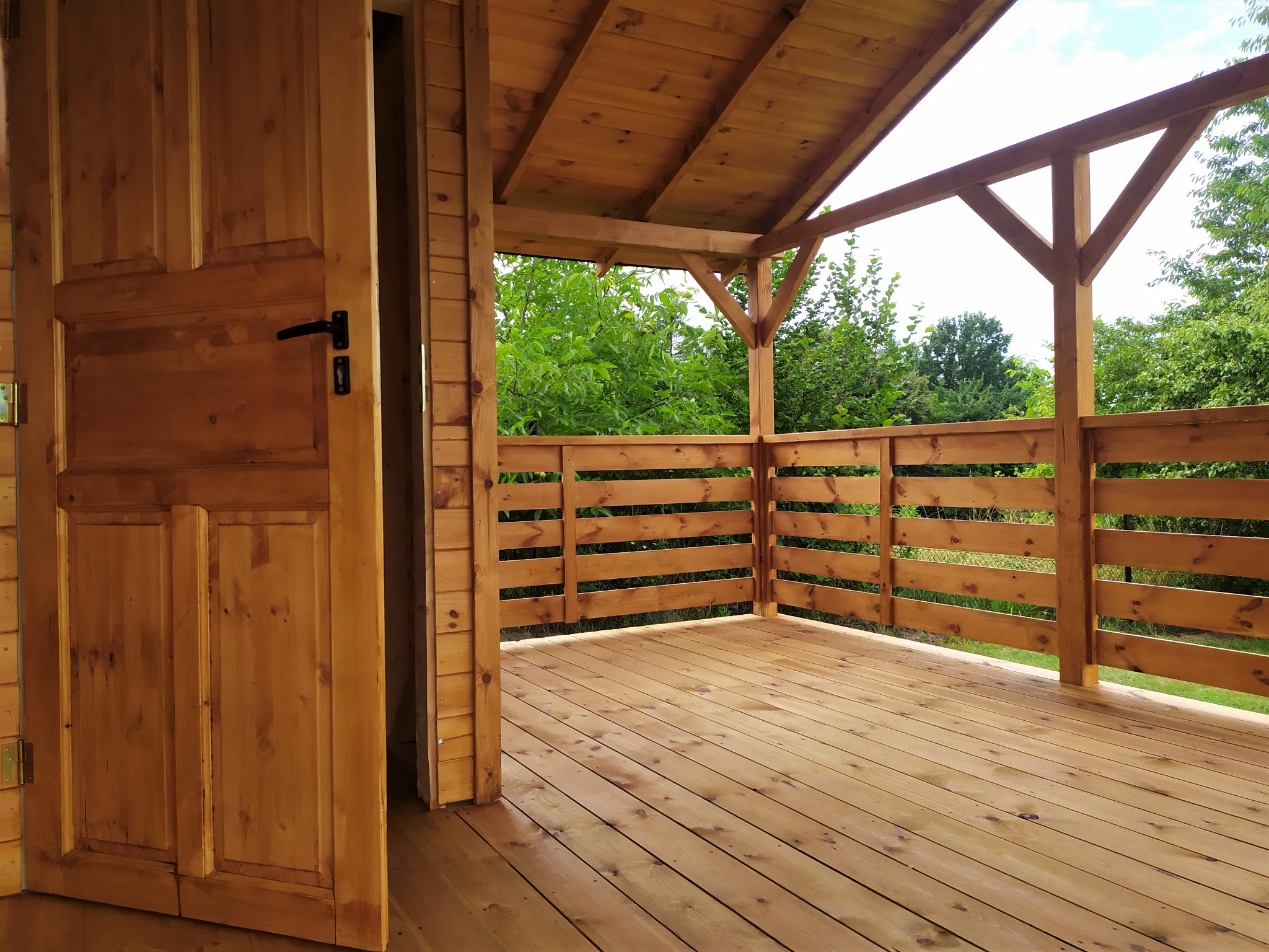 Drewniany taras z balustradą i otwartymi drzwiami, widok na zieleń ogrodu w tle.