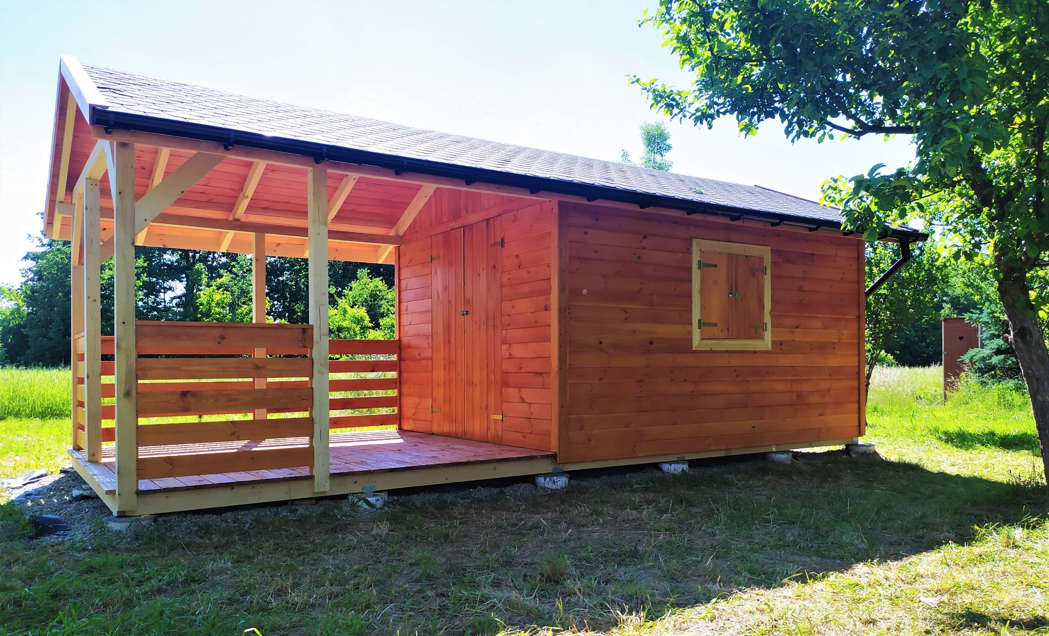 Drewniana konstrukcja z zadaszeniem i małą werandą, pomalowana na pomarańczowo-brązowy kolor, osadzona na betonowych bloczkach, z widocznym oknem z okiennicą i rynną, na tle zielonego trawnika i drzew