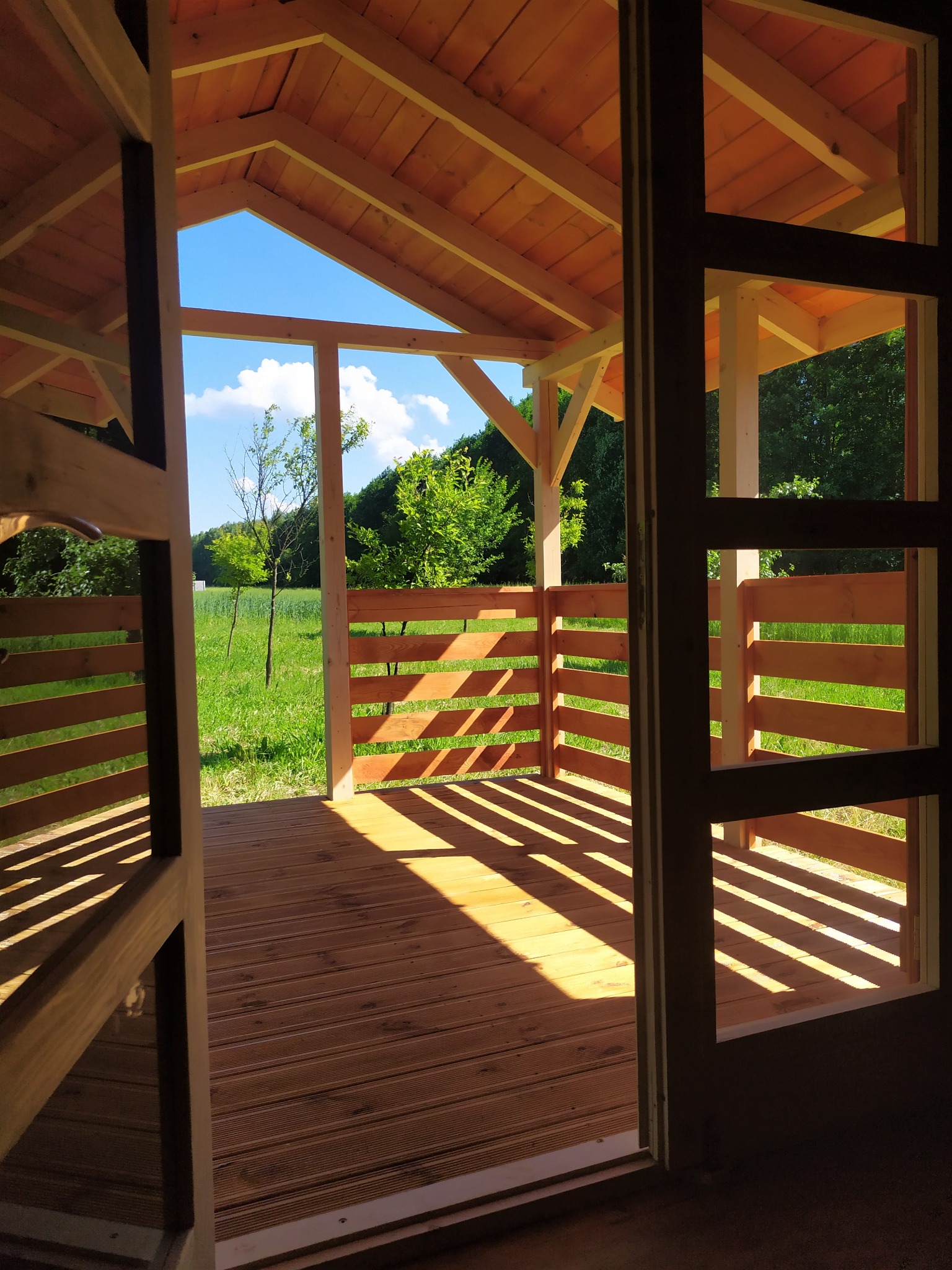 Wnętrze drewnianej altany szkieletowej z widokiem na zieloną łąkę i drzewa w słoneczny dzień, widoczne cienie na podłodze z desek.