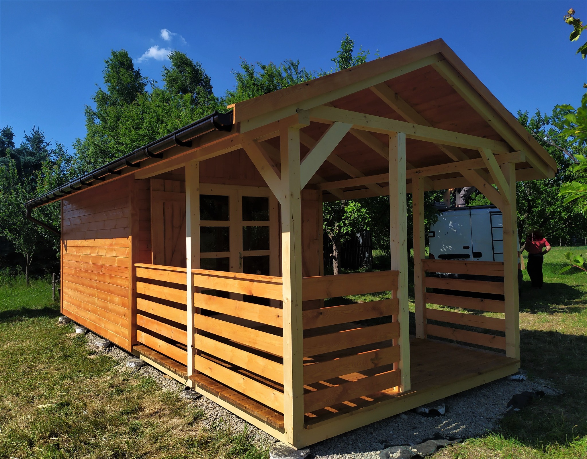 Surowy, drewniany domek letniskowy z przeszkloną ścianą boczną i otwartą werandą, częściowo wykończony, z pracownikiem na dachu samochodu w tle.