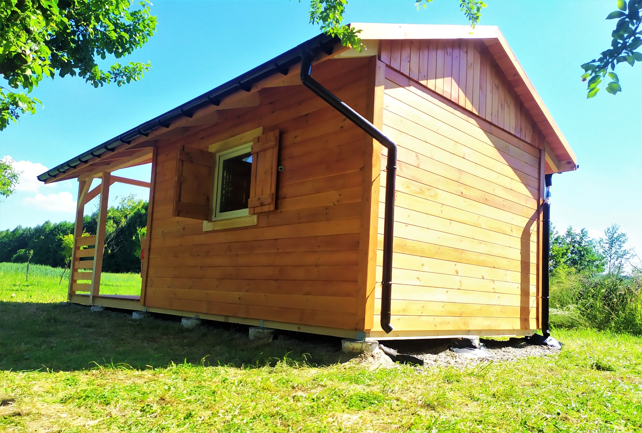 Niewielki, parterowy budynek z jasnego drewna z ciemnymi rynnami, z oknem z okiennicami, na betonowych bloczkach, otoczony zielonym trawnikiem i drzewami w tle, widziany w słoneczny dzień.