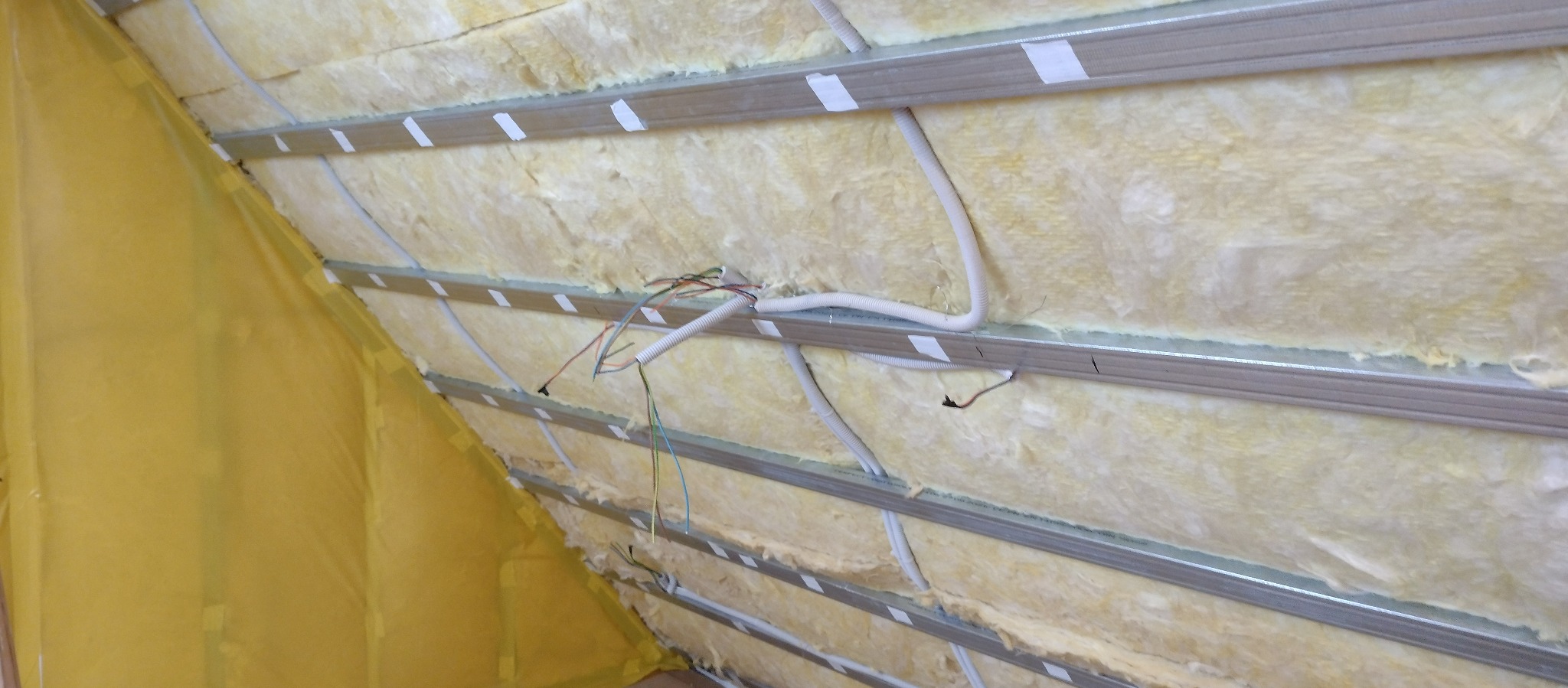 Ocieplenie poddasza wełną mineralną, widoczne profile metalowe, kable elektryczne w peszlach i folia paroizolacyjna przy ścianie szczytowej.