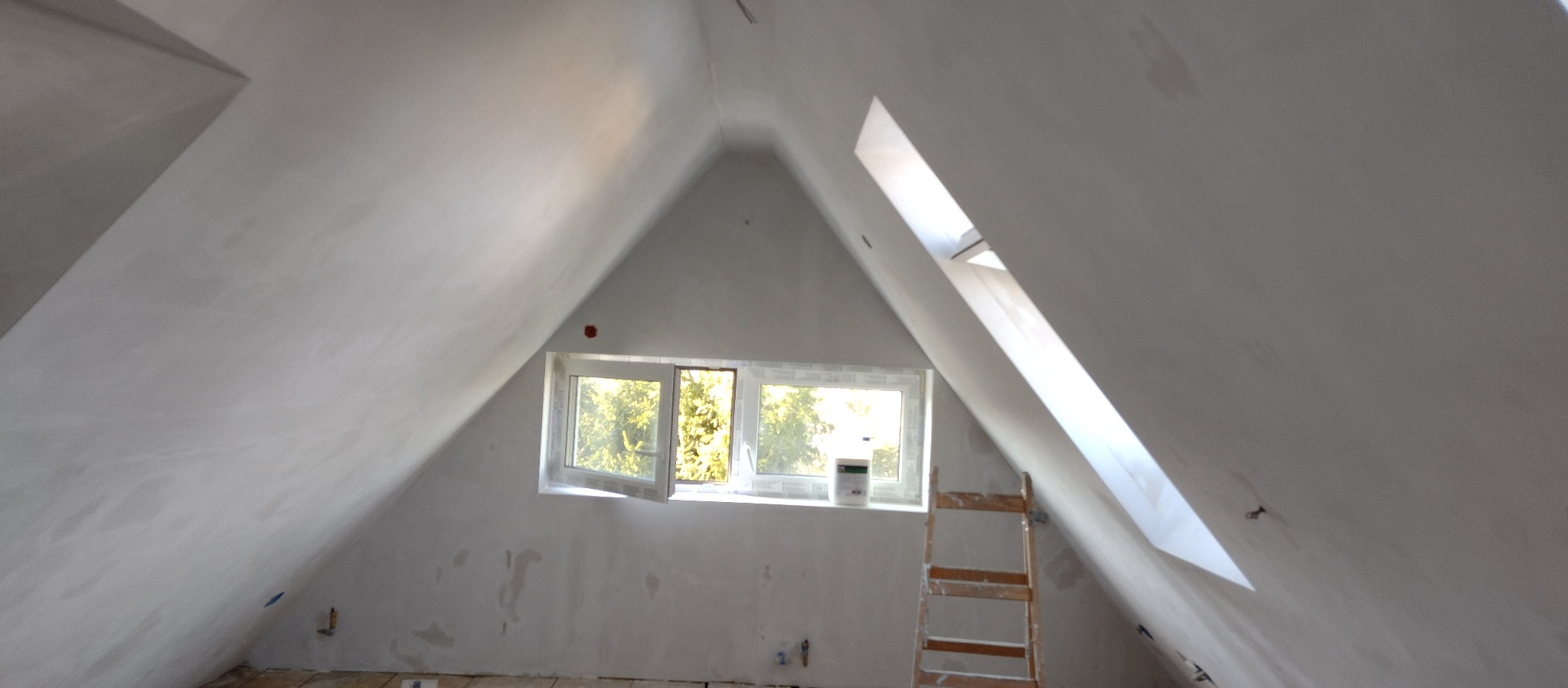Poddasze w trakcie remontu, ściany i sufit po szpachlowaniu, widoczne okno dachowe i drabina, ślady po szpachli na ścianach.