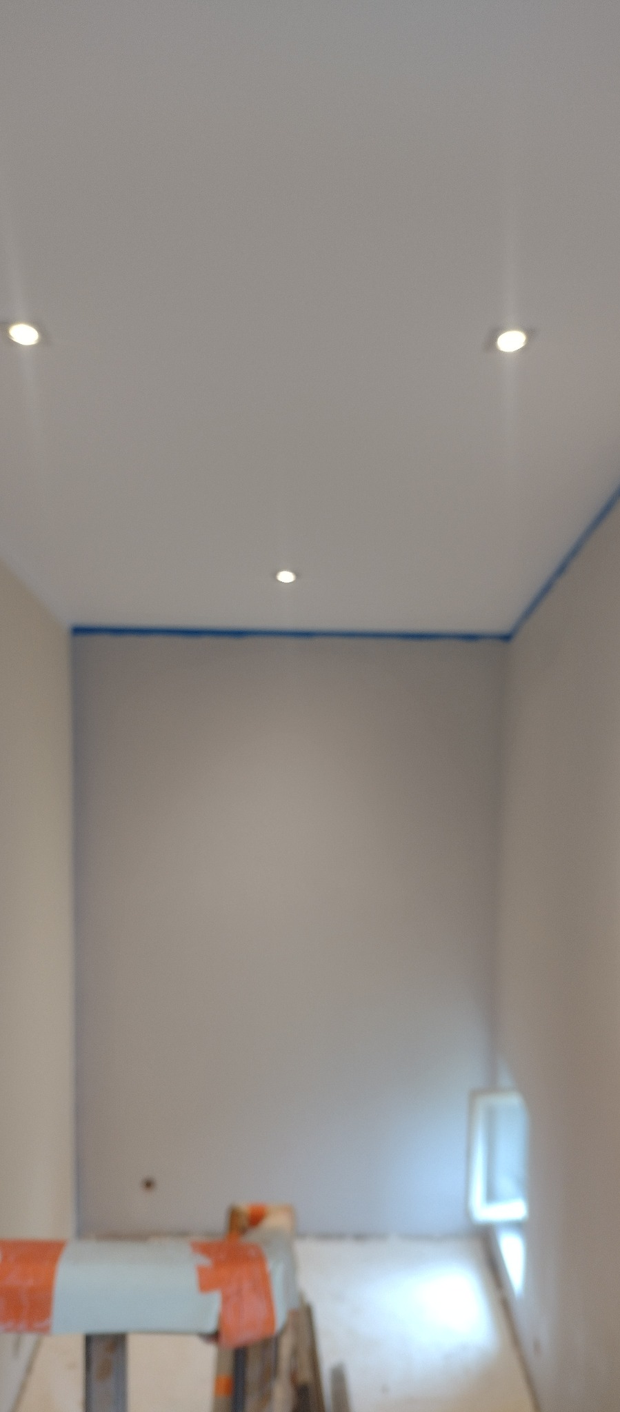 Wnętrze pokoju w trakcie prac przygotowawczych: ściany zabezpieczone taśmą malarską, sufit z punktowym oświetleniem, widoczny fragment rusztowania malarskiego.