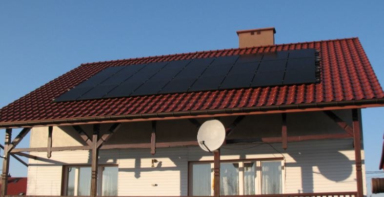 Panele słoneczne na dachu domu z czerwonej dachówki, widoczna antena satelitarna i fragment werandy.