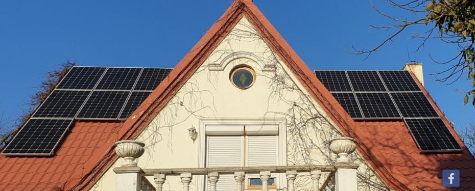 Panele słoneczne na dachu domu z czerwoną dachówką, pnącza na elewacji i okrągłe okno na poddaszu.