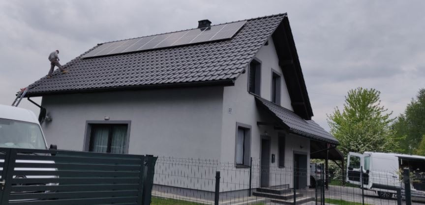Montaż paneli fotowoltaicznych na dachu dwuspadowym szarego domu jednorodzinnego; pracownik na drabinie, widoczne dwa samochody dostawcze.