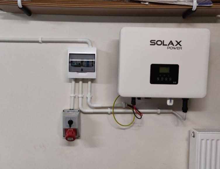 Biały inwerter Solax Power X3 zamontowany na ścianie obok skrzynki elektrycznej i wyłącznika bezpieczeństwa, widoczne okablowanie w białych rurkach.
